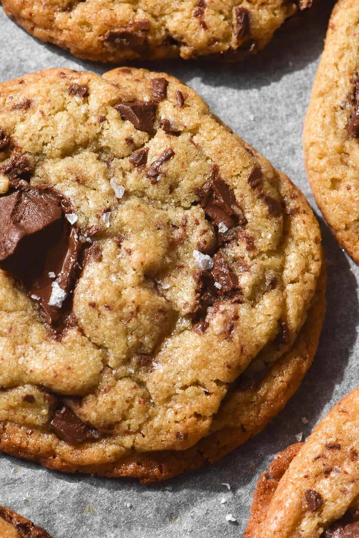 An aerial close up image of gluten free buckwheat choc chip cookies atop a lined baking sheet. The cookie is golden brown, studded with melty chocolate and topped with a scant sprinkle of sea salt flakes.