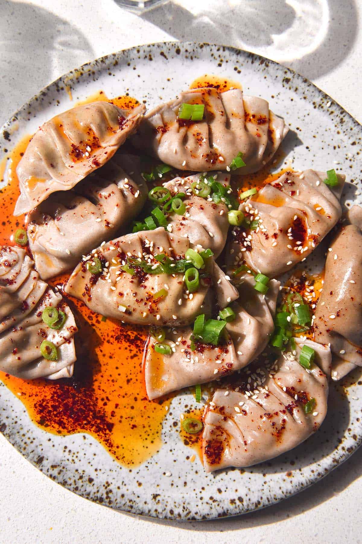 A brightly lit aerial image of gluten free dumplings on a white speckled ceramic plate atop a white stone bench. The dumplings are doused in bright red chilli oil and topped with spring onion greens and sesame seeds. 