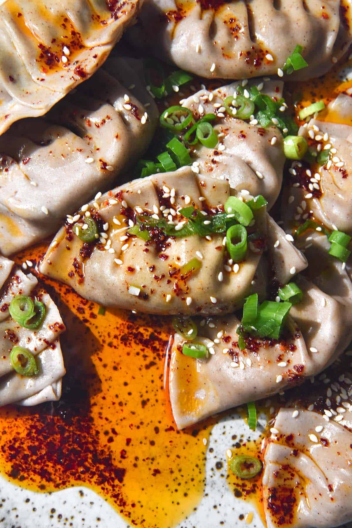 A brightly lit aerial close up image of gluten free dumplings on a white speckled ceramic plate atop a white stone bench. The dumplings are doused in bright red chilli oil and topped with spring onion greens and sesame seeds. 