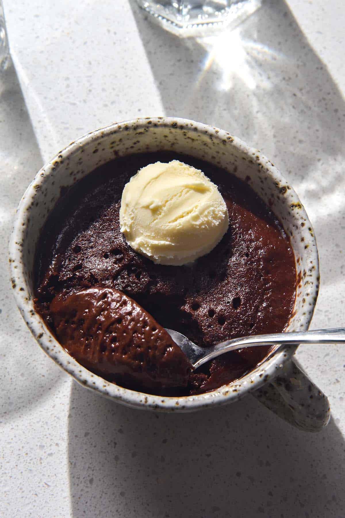 An aerial image of a gluten free vegan chocolate mug cake on a brightly lit white stone benchtop. The mug cake is topped with a scoop of vanilla ice cream and the mug is framed by light shining through two water glasses. 