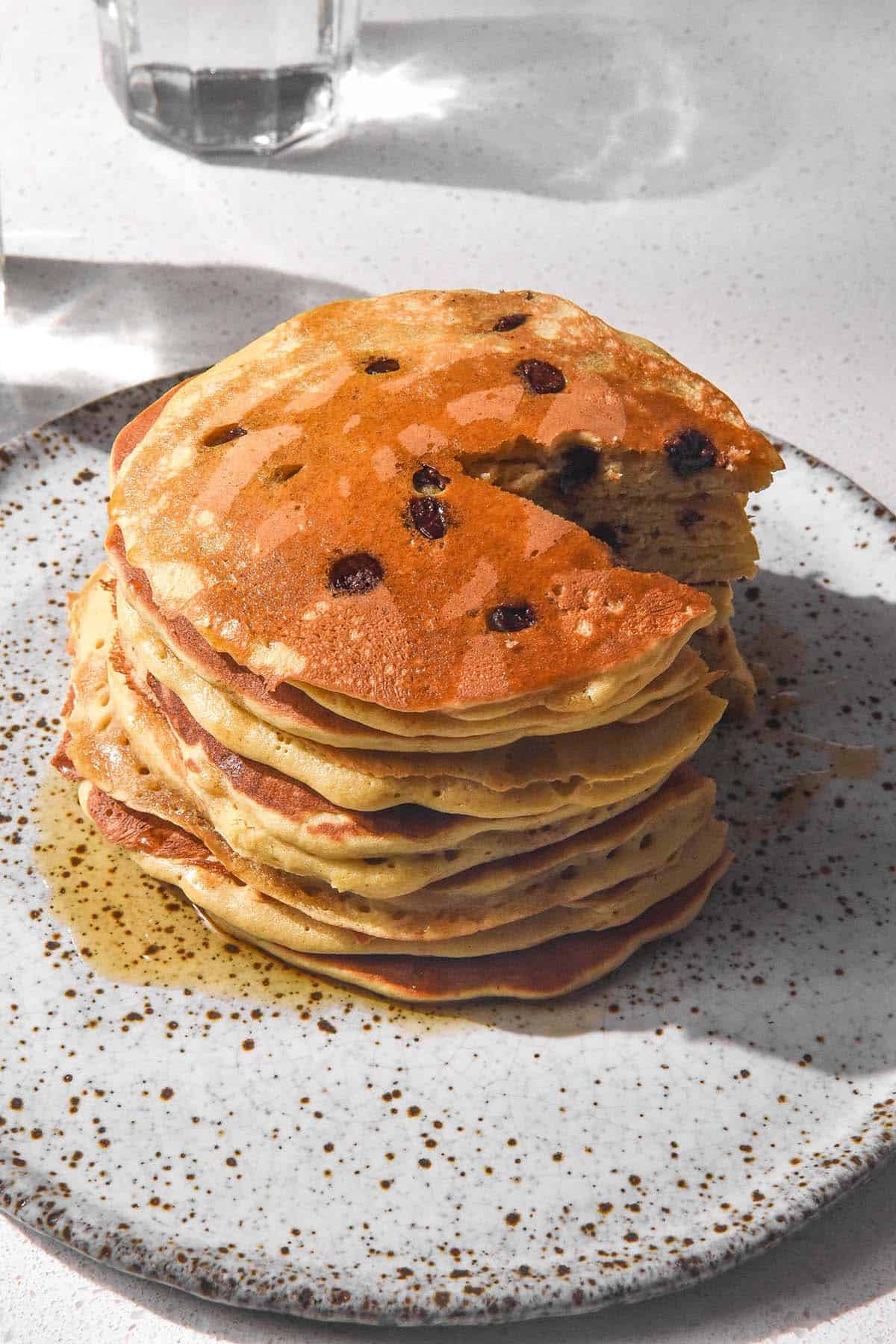 A brightly lit image of a stack of buckwheat flour choc chip pancakes. The slice sits on a white speckled ceramic plate atop a white stone benchtop. Two glasses of water cast shadows onto the benchtop in the background. The pancake stack has been sliced and chocolate chips stud the golden brown pancakes. 