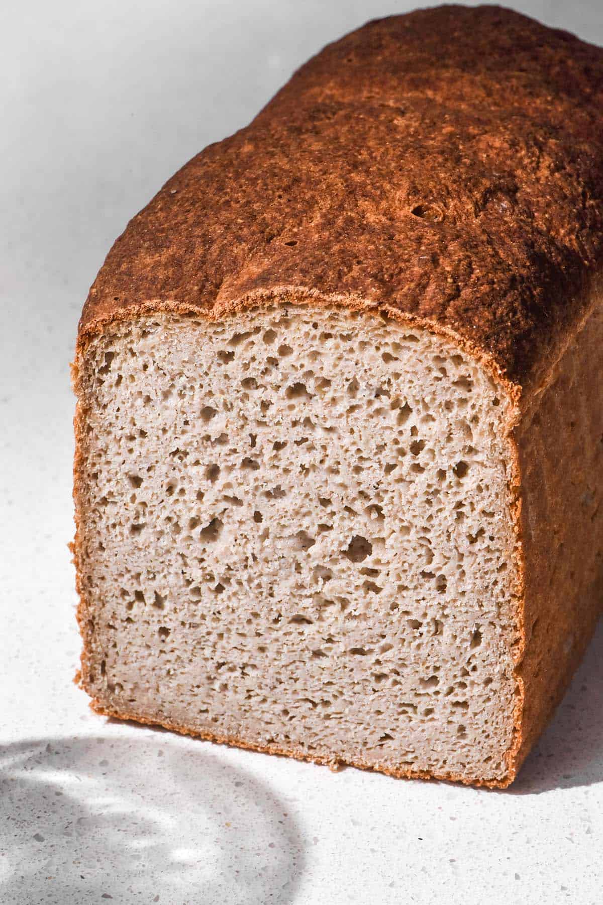 A side on, brightly lit image of a loaf of 100% buckwheat flour bread. The loaf has been sliced to reveal a soft crumb inside. It sits on a white stone bench top and the shadow of a water glass contours the bottom left of the image. 