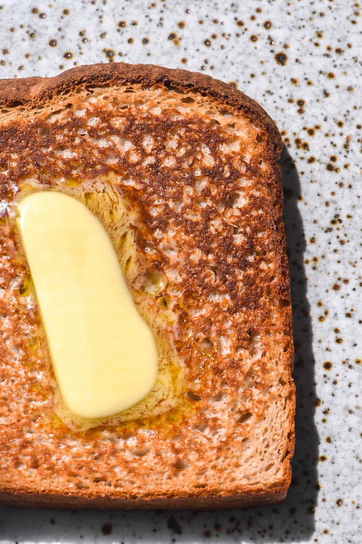 A brightly lit macro image of a slice of toasted buckwheat sourdough on a white speckled ceramic plate. The toast is topped with melting butter. 