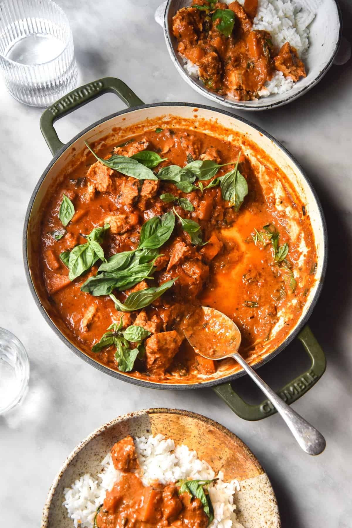 An aerial image of a low FODMAP Thai red curry in a skillet and topped with Thai basil leaves. The curry sits atop a white marble table and is surrounded by other bowls of curry. 