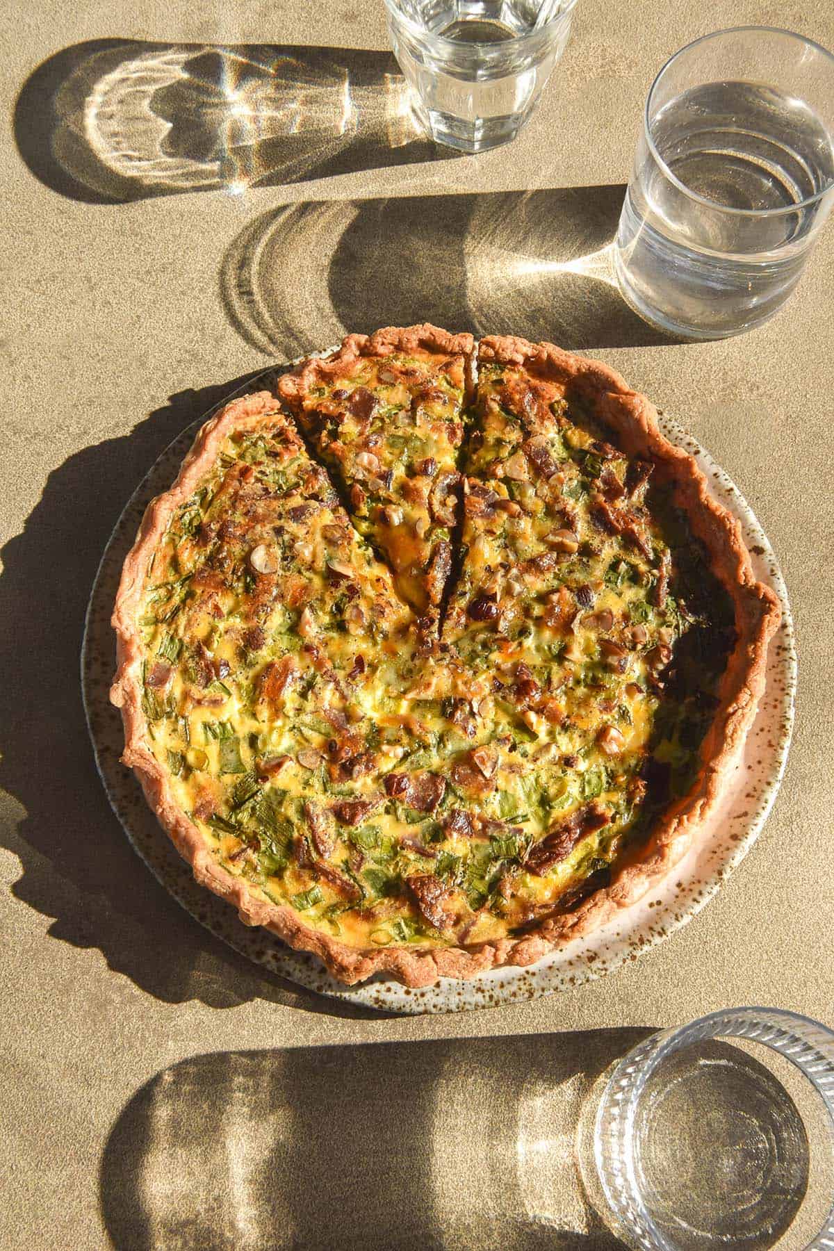 An aerial sunlit image of a gluten free quiche on a white speckled ceramic plate atop a stone table. Three sunlit glasses of water surround the quiche. 