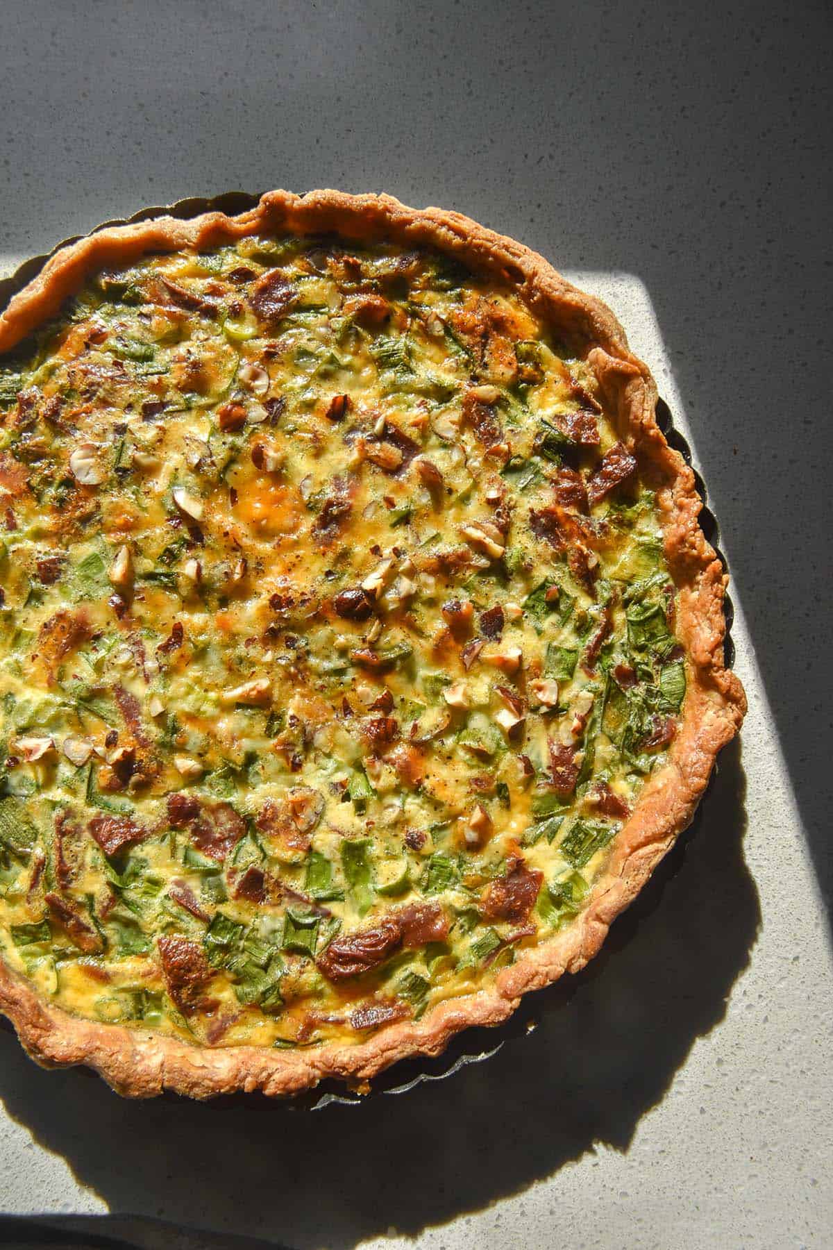 An aerial image of a gluten free quiche filled with spring onion greens and vegetarian bacon. The quiche sits atop a white speckled ceramic plate on a white stone benchtop.