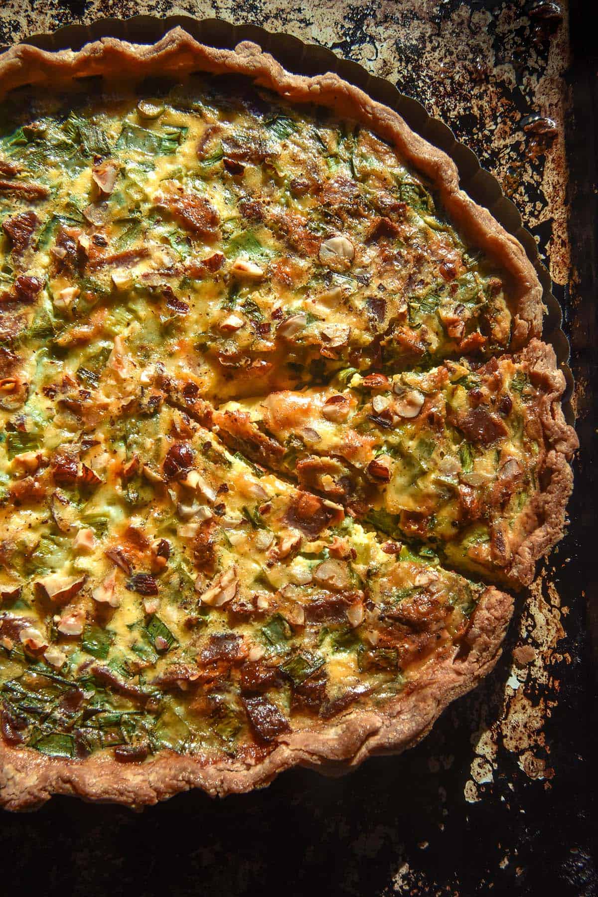 An aerial image of a gluten free vegetarian bacon and spring onion quiche on a mottled dark baking tray. 