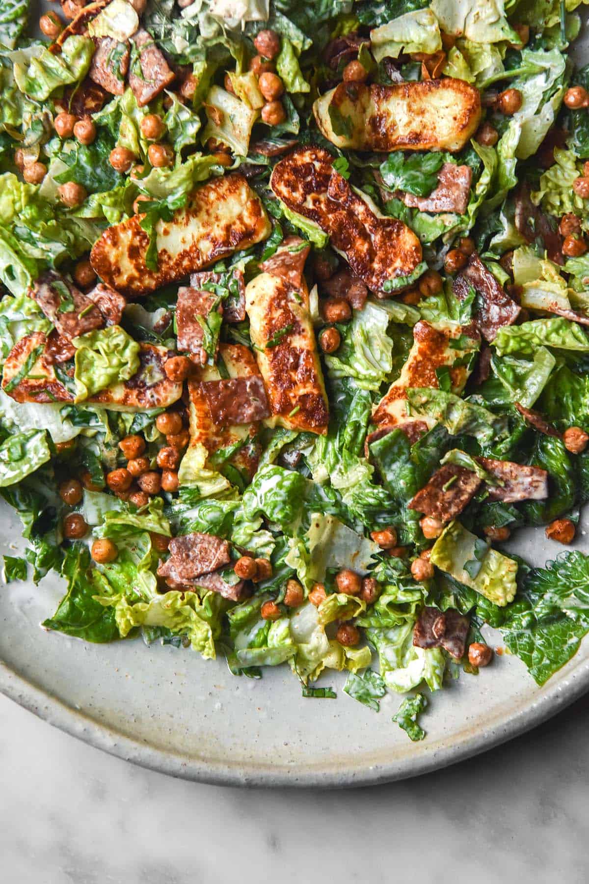 An aerial close up image of a lettuce, haloumi, crispy chickpea and vegetarian bacon salad. The salad sits on a white ceramic serving platter atop a white marble table