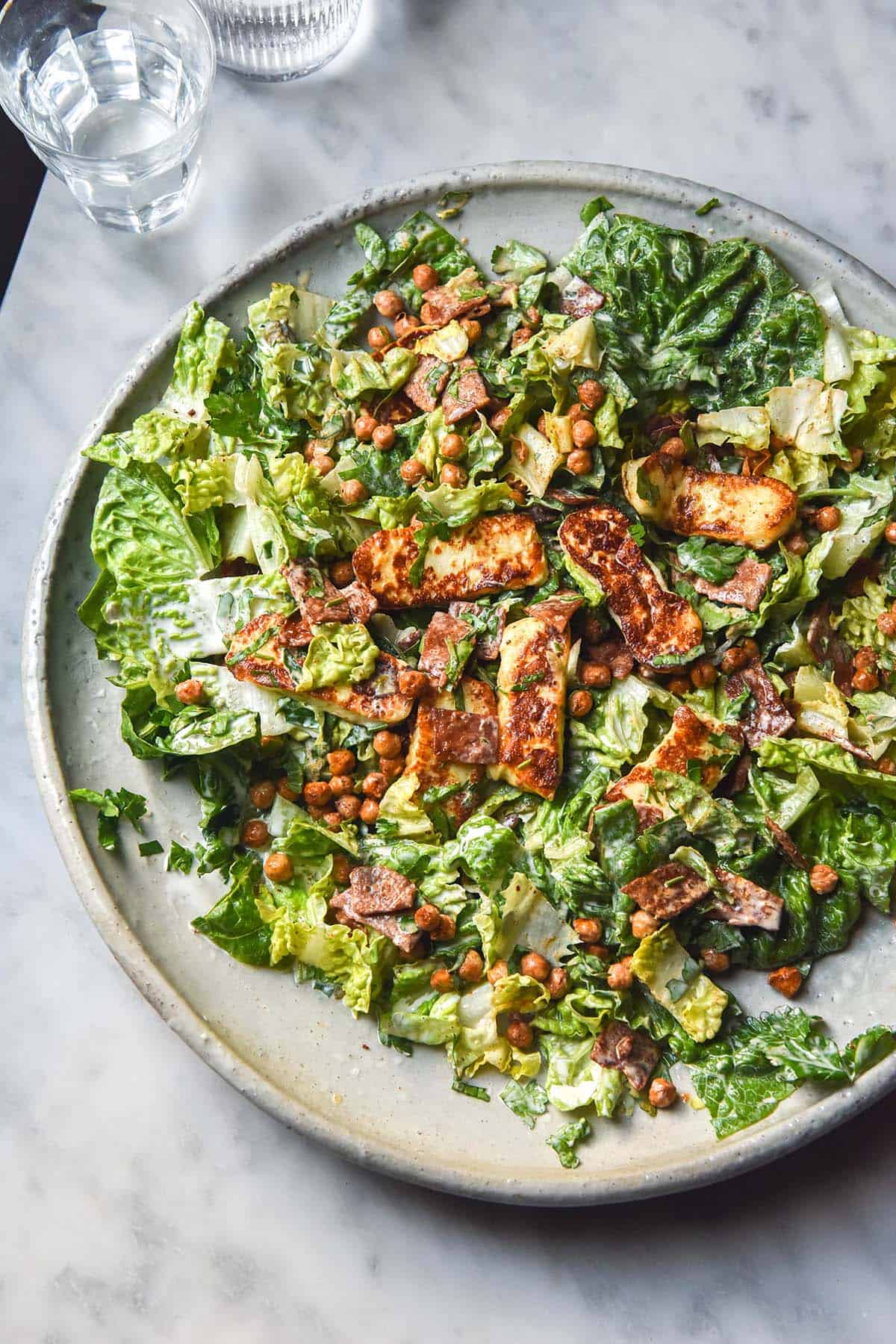 An aerial image of a lettuce, haloumi, crispy chickpea and vegetarian bacon salad. The salad sits on a white ceramic serving platter atop a white marble table. Two glasses of water sit to the top left of the image. 