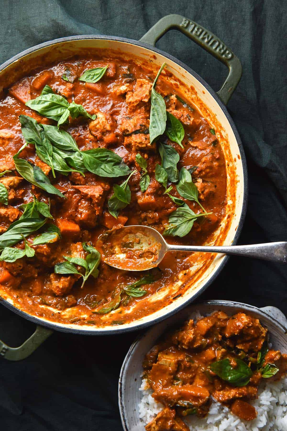 An aerial image of a skillet filled with low FODMAP Thai red curry which is topped with Thai Basil. The skillet sits atop an olive green linen tablecloth and a bowl of rice and curry sits to the bottom right of the skillet