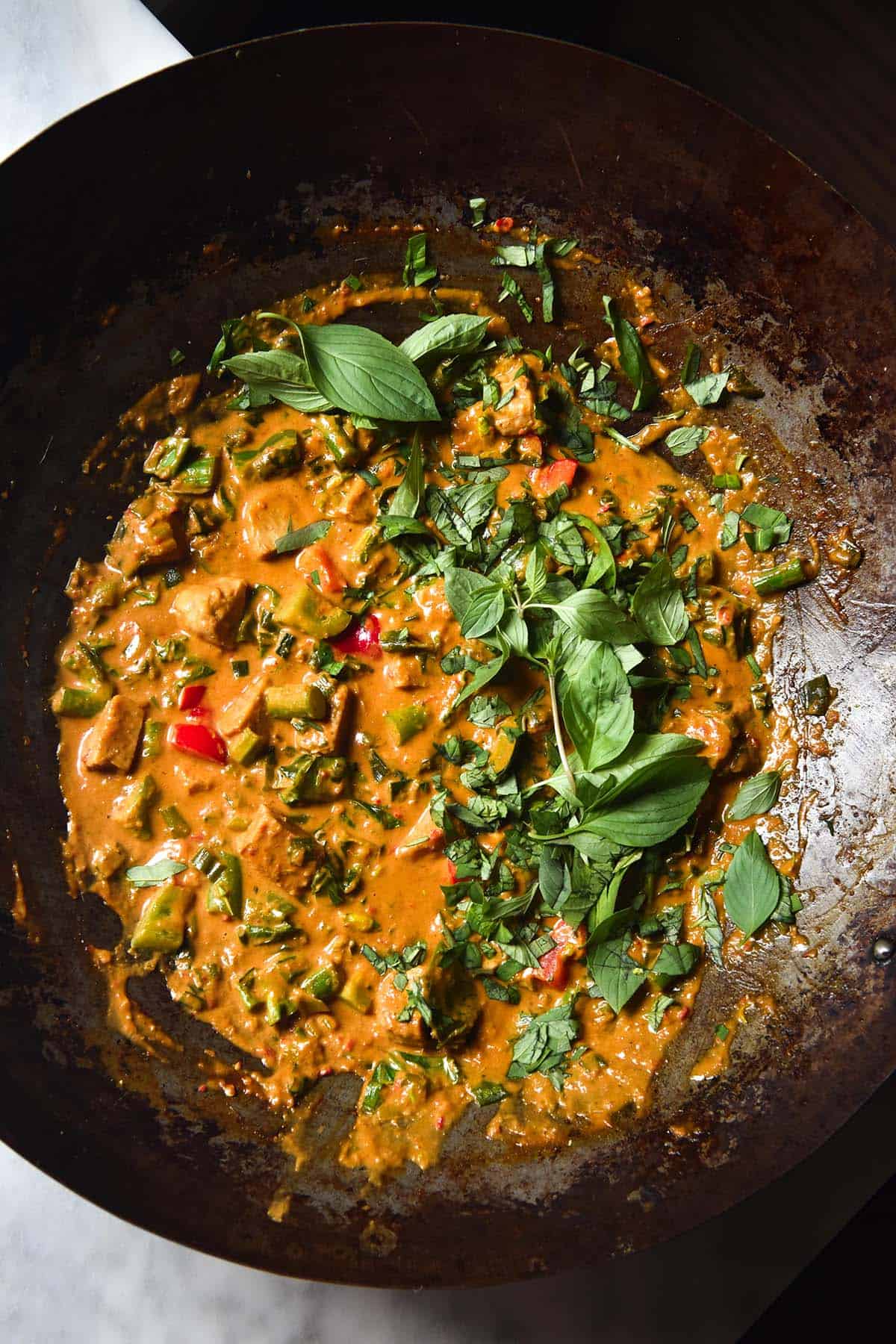 An aerial image of vegan low FODMAP Thai red curry in a mottled skillet. The curry is topped with casually strewn Thai basil leaves and sits on a white marble table.