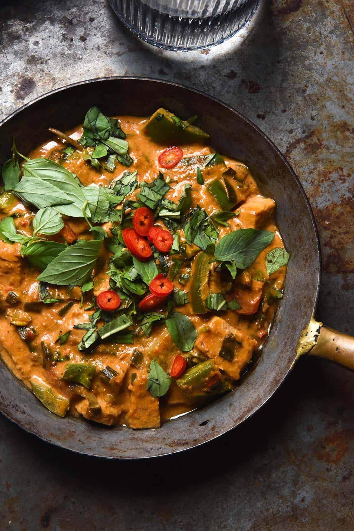 An aerial image of a mini skillet filled with low FODMAP Thai red curry. The skillet sits on a dark steel backdrop and a glass of water sits to the top of the image. The curry is topped with Thai basil leaves and slices of red chilli