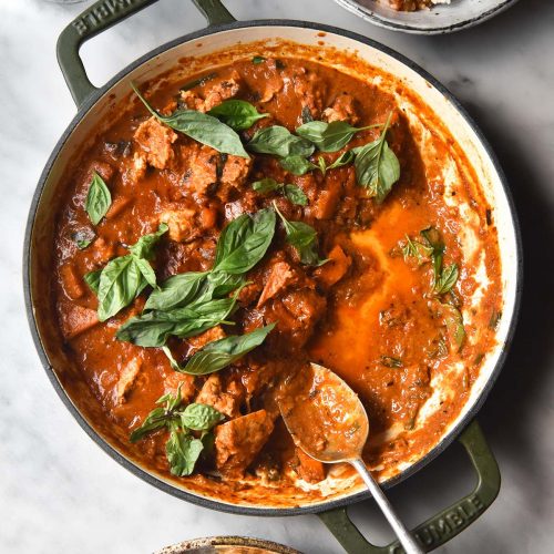 An aerial image of a skillet filled with low FODMAP Thai red curry on a white marble table. The skillet is surrounded by two extra bowls of rice and curry and a glass of water sits to the top left of the image