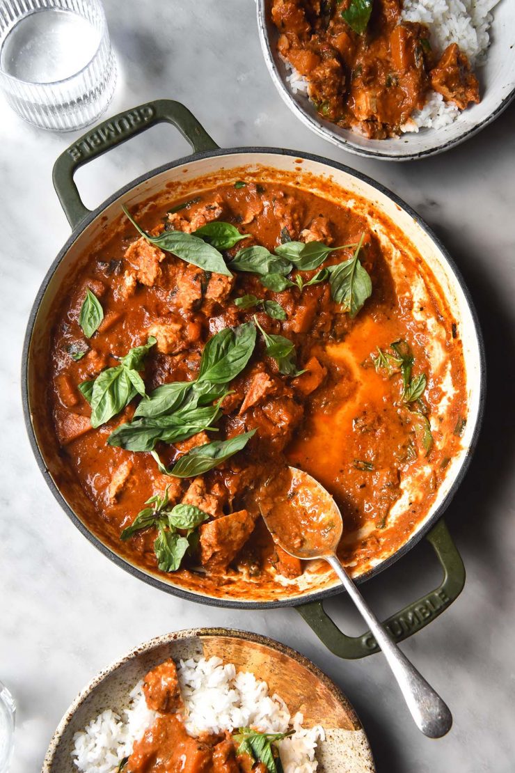 An aerial image of a skillet filled with low FODMAP Thai red curry on a white marble table. The skillet is surrounded by two extra bowls of rice and curry and a glass of water sits to the top left of the image