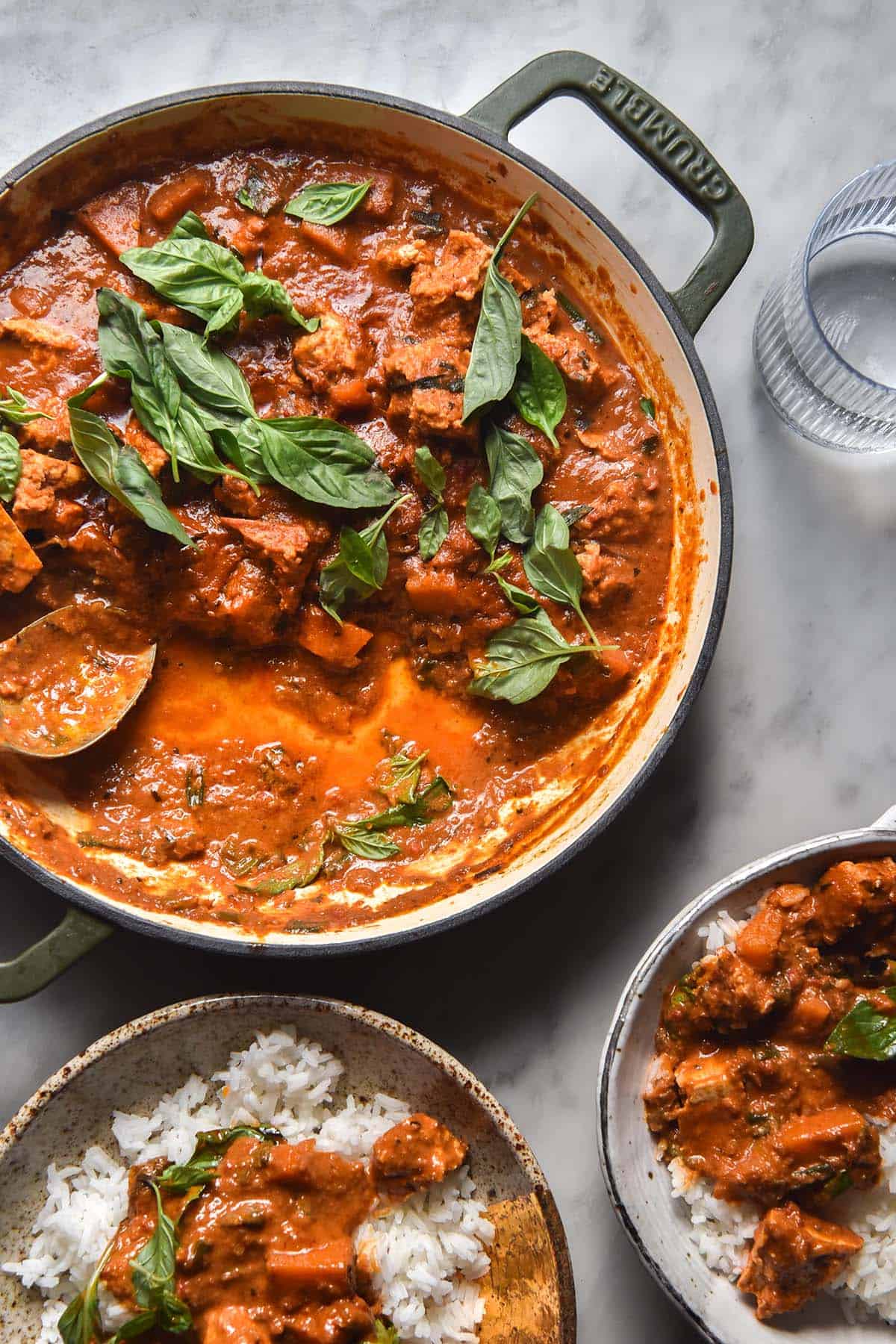 An aerial image of a skillet filled with vegan Thai red curry topped with Thai basil leaves. The skillet sits atop a white marble table and two bowls of rice and curry sit in the bottom of the image.