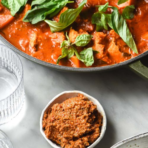 An aerial image of a small bowl of low FODMAP Thai red curry paste on a white marble table. The paste is surrounded by a white bowl of red, water glasses and an olive green skillet filled with Thai red curry.