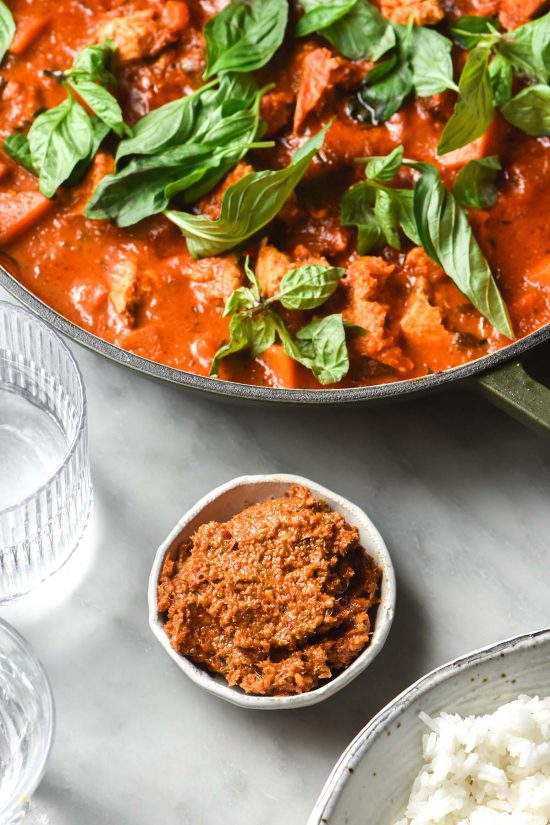 An aerial image of a small bowl of low FODMAP Thai red curry paste on a white marble table. The paste is surrounded by a white bowl of red, water glasses and an olive green skillet filled with Thai red curry.