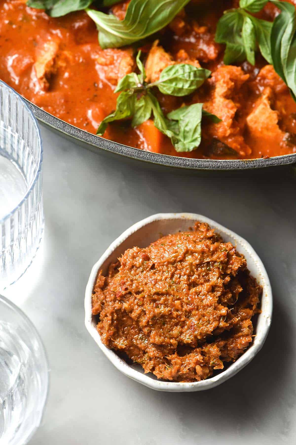 An aerial close up image of a small white bowl filled with low FODMAP Thai red curry paste. The bowl sits on a white marble table and is surrounded by water glasses and a skillet filled with Red Thai curry. 