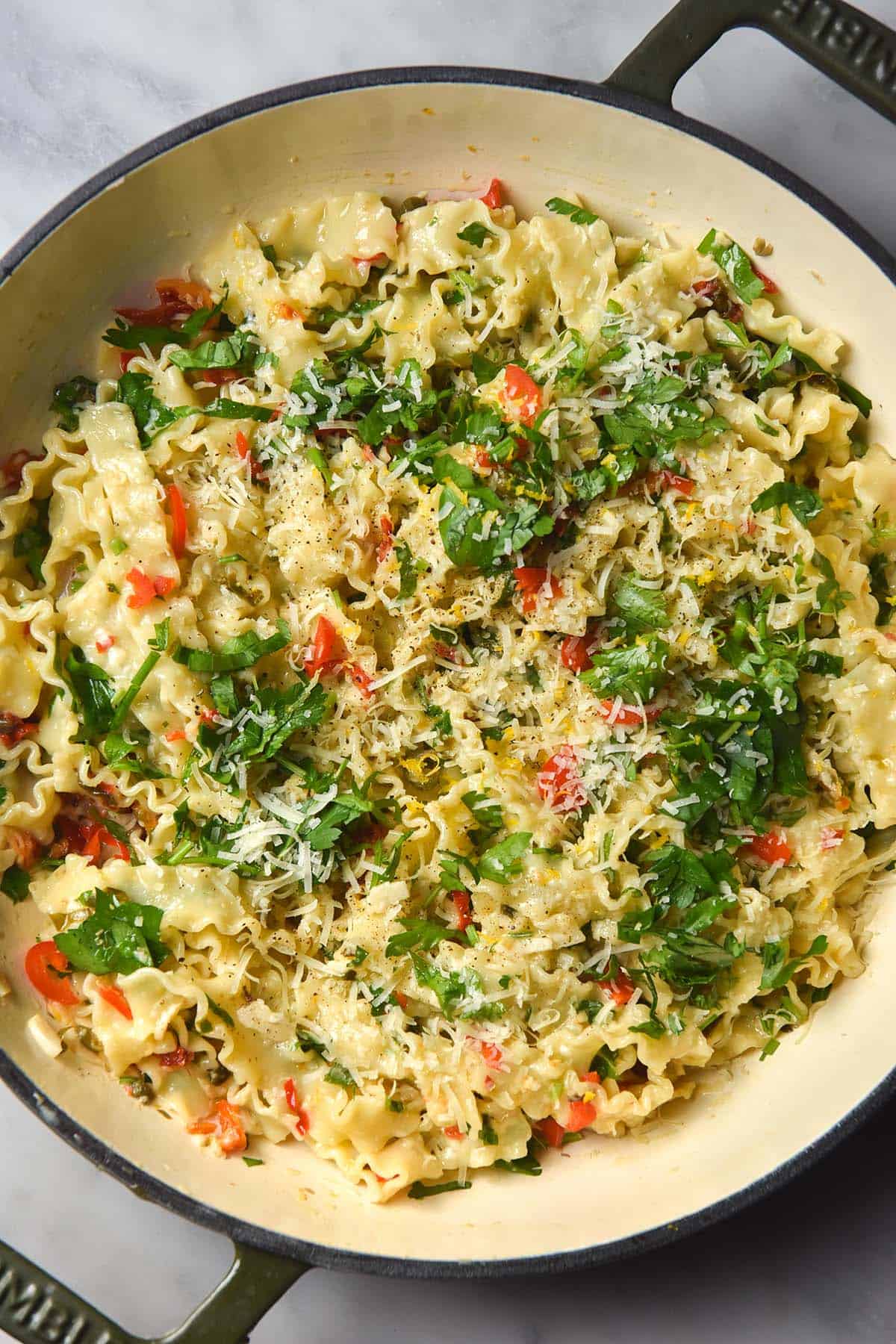 An aerial image of a low FODMAP pasta dish in a an olive green skillet atop a white marble table. The pasta is topped with parsley, parmesan and red chlli.