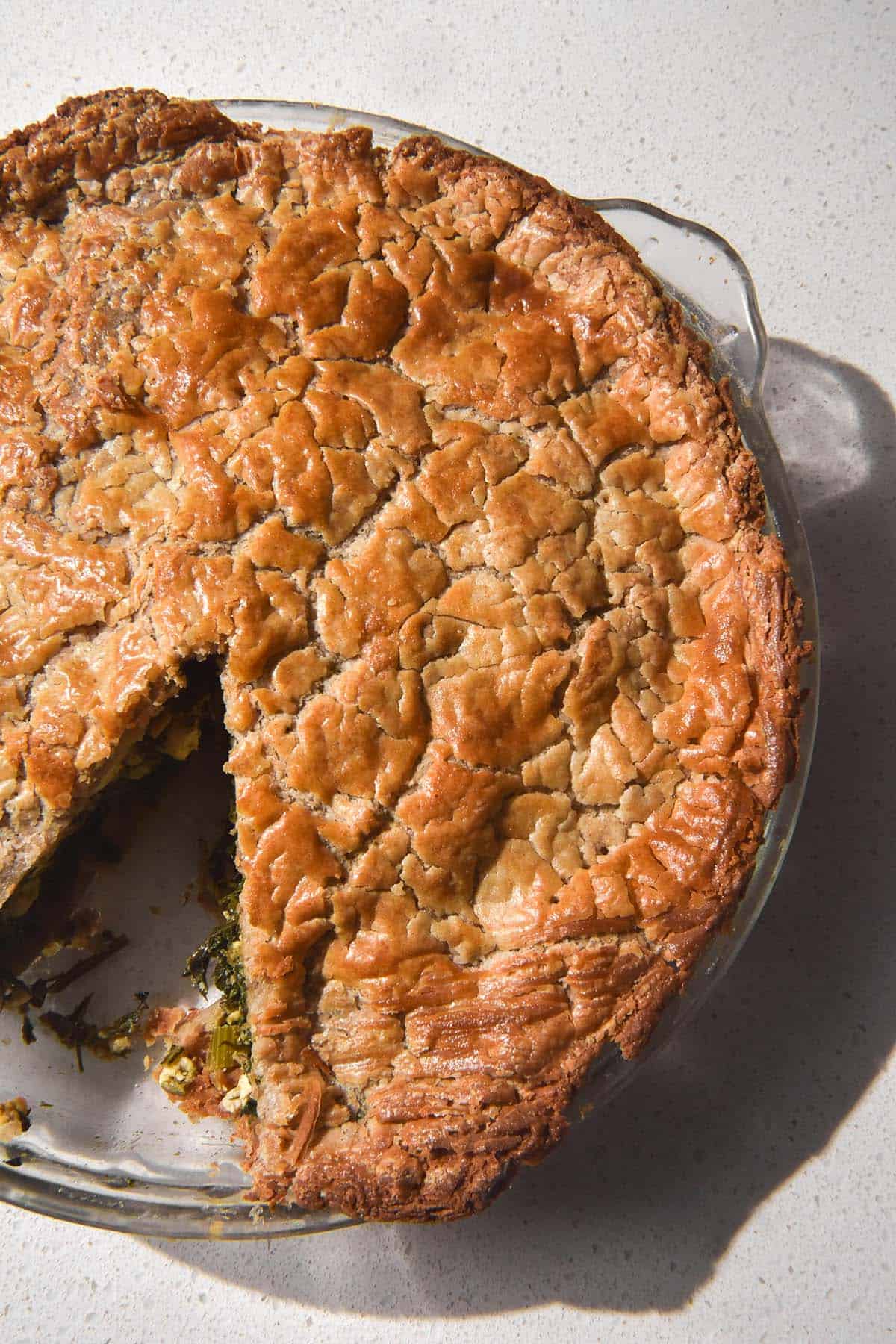 An aerial image of a kale and feta pie made with buckwheat puff pastry. The pie sits in a glass pie dish on a white stone bench in bright light.