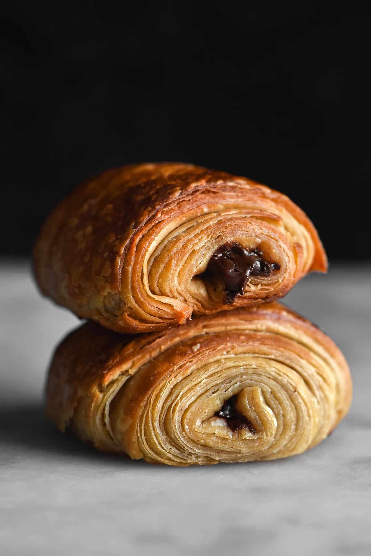A side on image of two gluten free pain au chocolat stacked on top of eachother. They are golden brown and chocolate oozes from the seam closest the camera. The pains au chocolat sit on a white marble table against a black backdrop. 