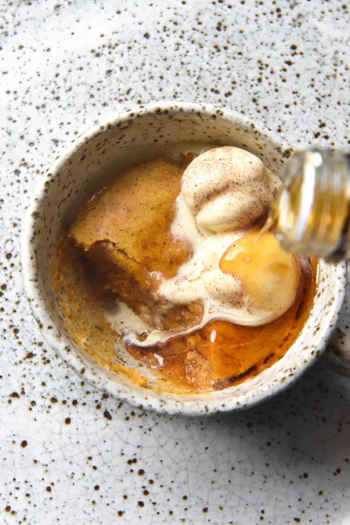 An aerial image of a gluten free pumpkin mug cake in a white speckled ceramic mug atop a white speckled ceramic plate. The mug cake is topped with melting vanilla ice cream and maple syrup. 
