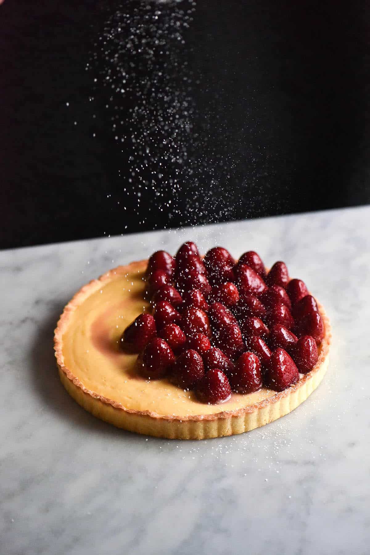 A side on view of a gluten free custard and berry tart on a white marble table against a black backdrop. The tart is topped with small upright strawberries and is being sprinkled with a dusting of icing sugar.