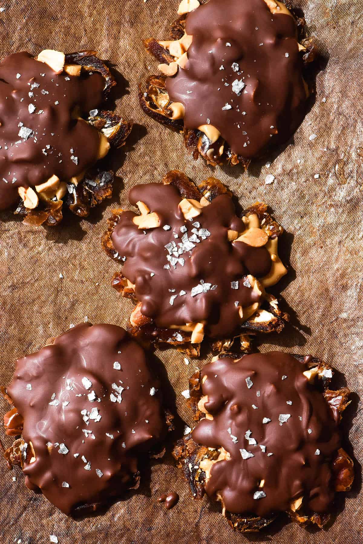 A moody aerial image of five dark bark bites on a lined baking tray. The dates are covered in peanut butter, peanuts, dark chocolate and sea salt flakes. 