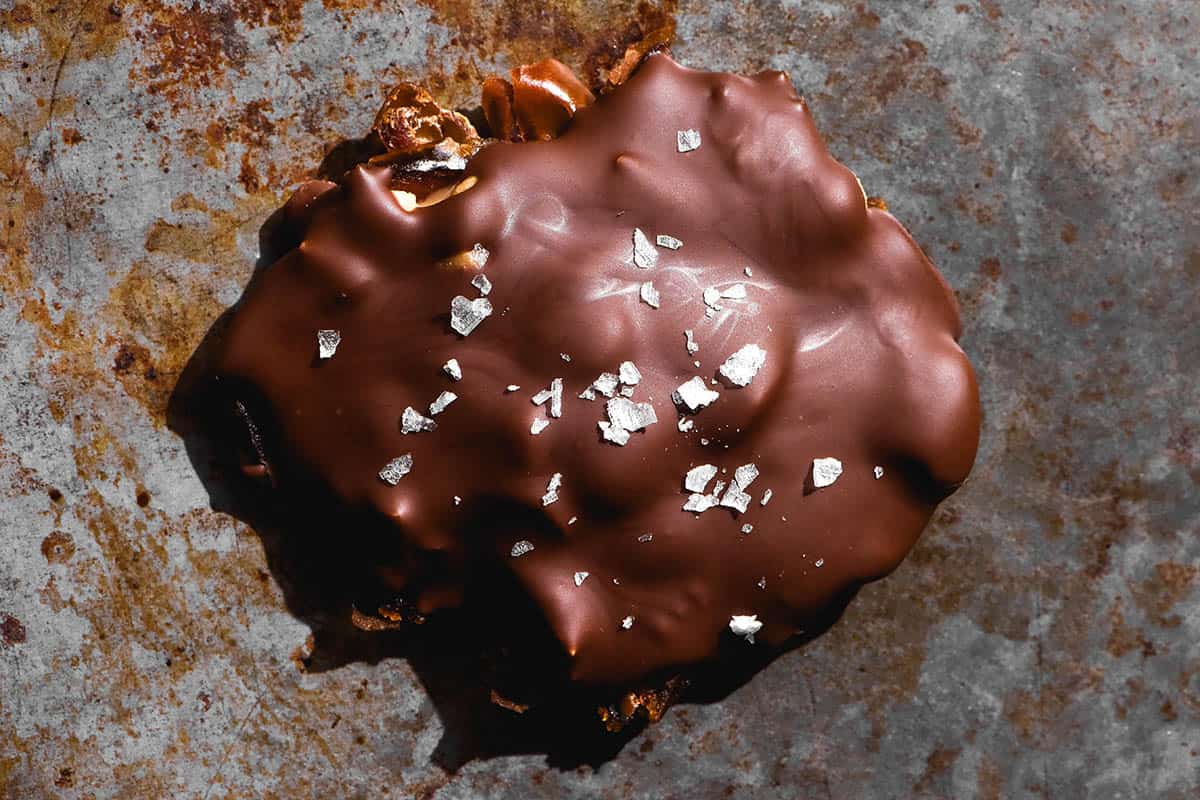 An aerial close up image of a date bark bite on a mottled steel backdrop. The date is topped with chocolate and sea salt flakes. 