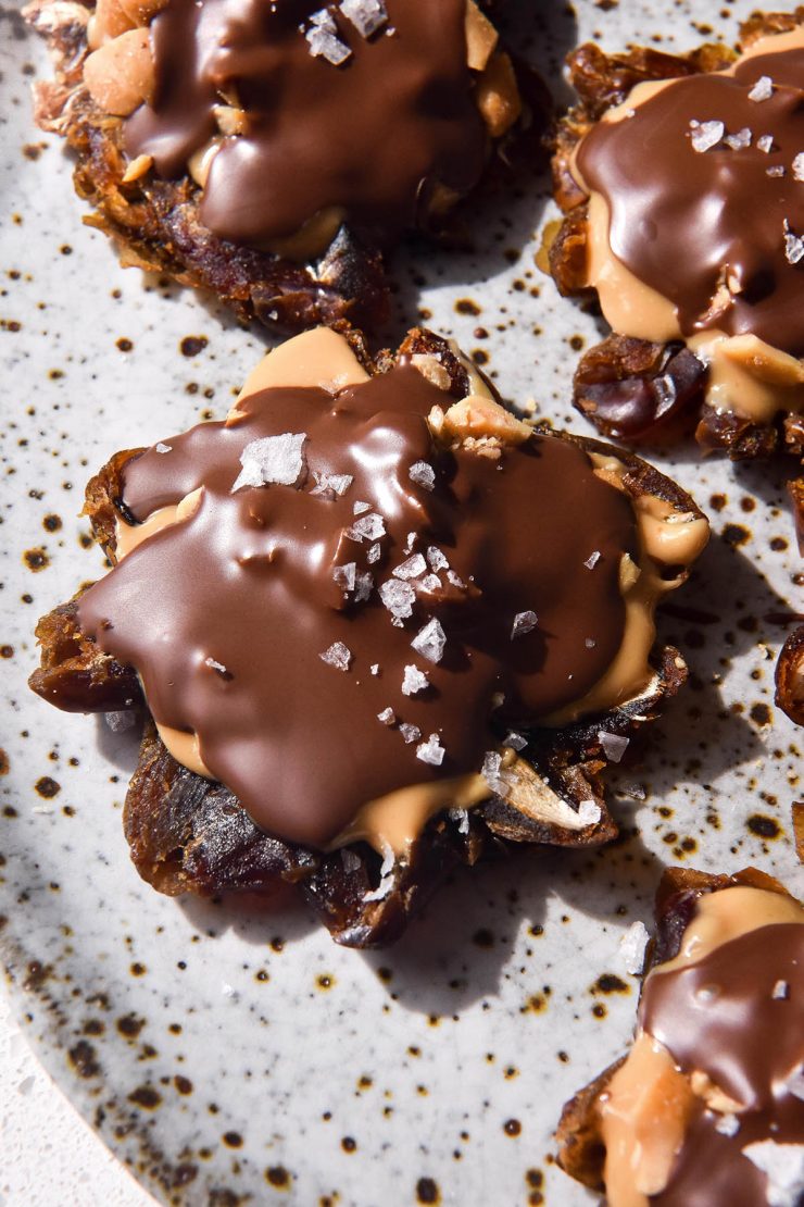 A brightly lit image of date bark bites with peanuts, butter butter, a chocolate coating and a sprinkle of sea salt flakes. The bites sit on a white speckled ceramic plate atop a white bench top.