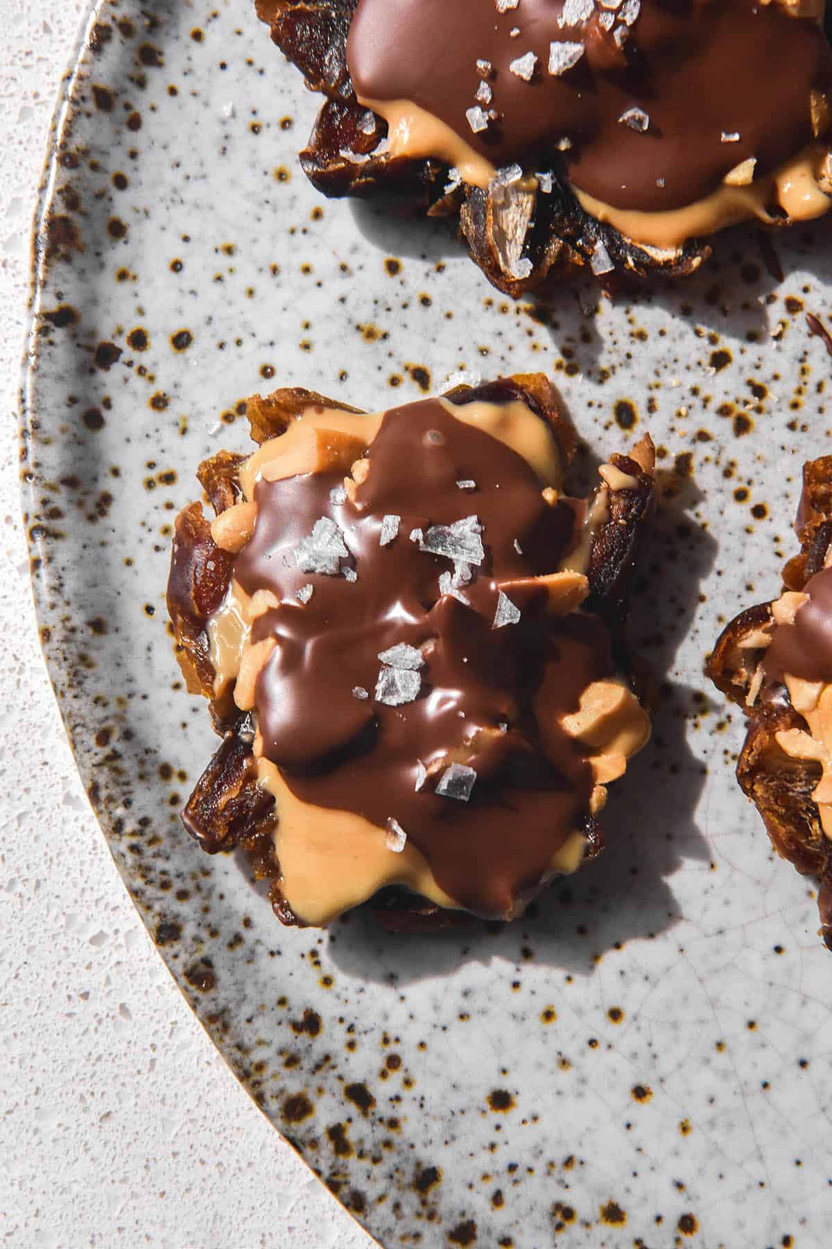 An aerial macro image of date bark bites topped with peanuts, peanut butter and chocolate then finished with sea salt flakes. The bites sit on a white speckled ceramic plate atop a white bench top. 