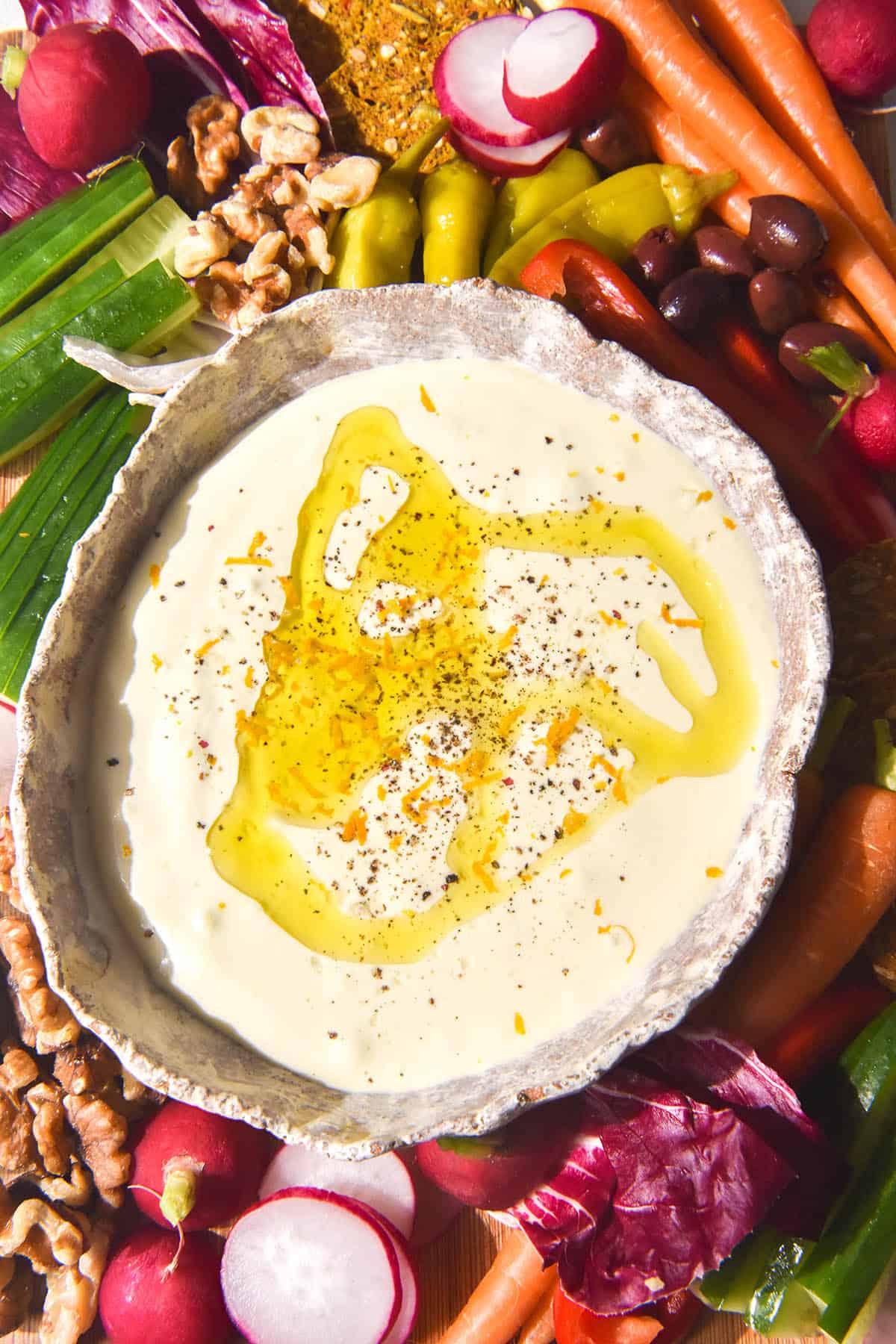 A brightly lit aerial image of a white ceramic bowl filled with garlic dip which is topped with a drizzle of oil, salt, pepper and lemon zest. The bowl is surrounded by fresh crudites, nuts and olives.