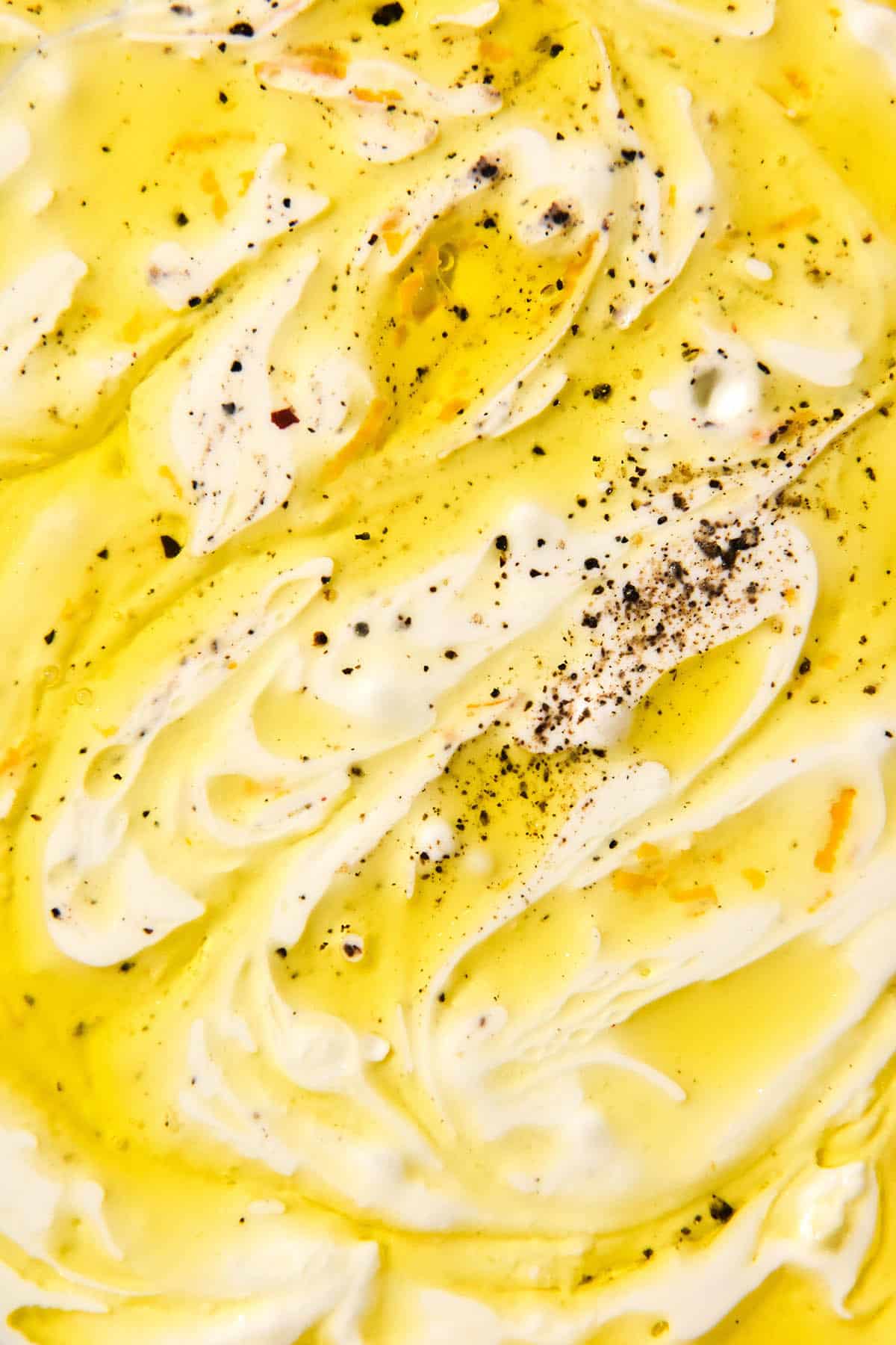 An aerial macro image of the top of a bowl of garlic dip splashed with olive oil and seasoned with salt, pepper and lemon zest