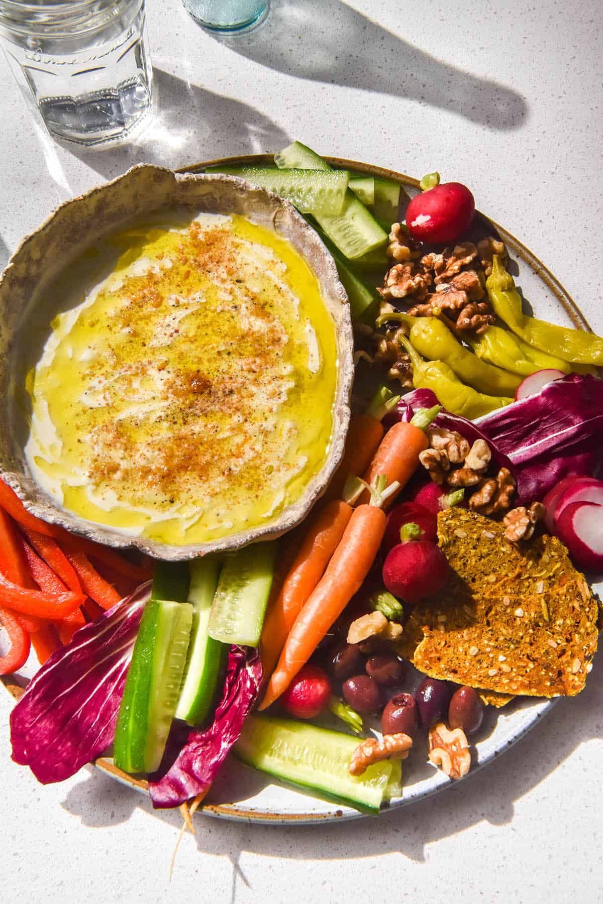 An aerial image of a white ceramic bowl filled with low FODMAP garlic dip atop a plate of rainbow coloured crudites. The plate sits atop a white stone bench and water glasses sit to the top right of the image.