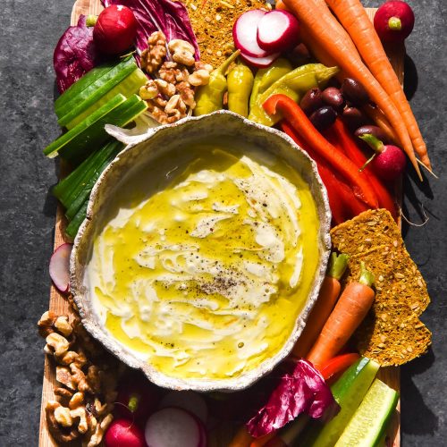 An aerial image of a white ceramic bowl filled with low FODMAP garlic dip on a crudite board. The board sits atop a dark blue backdrop and water glasses sit to the top of the image.