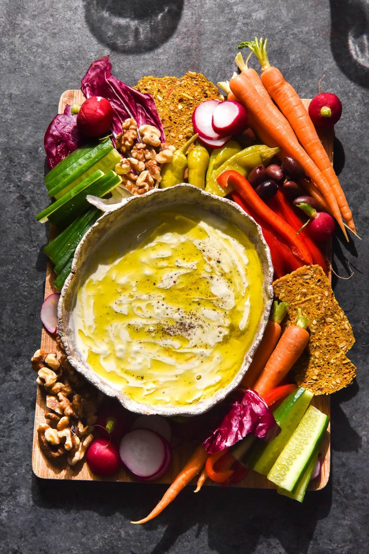 An aerial image of a white ceramic bowl filled with low FODMAP garlic dip on a crudite board. The board sits atop a dark blue backdrop and water glasses sit to the top of the image.