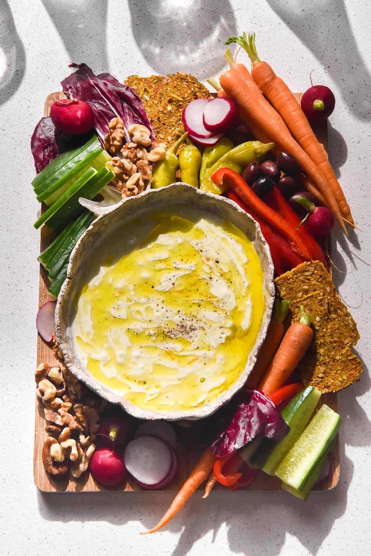 A brightly lit aerial image of low FODMAP garlic dip in a white ceramic bowl atop a wooden board of rainbow coloured crudites. The board sits on a white stone bench top with the shadows of water glasses in the top of the image.