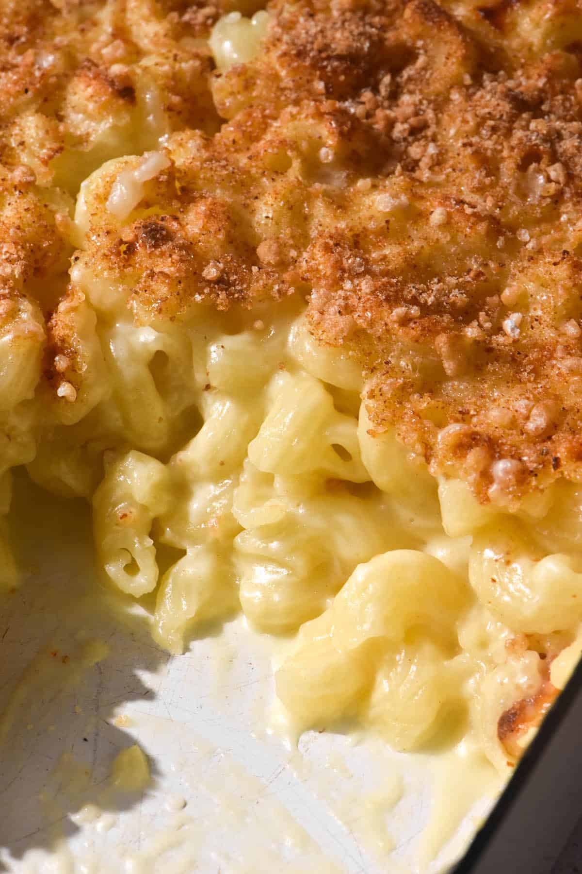 A close up of a mac and cheese bake in a white baking dish. Some of the bake has been removed, revealing the creamy mac and cheese underneath the golden brown topping.