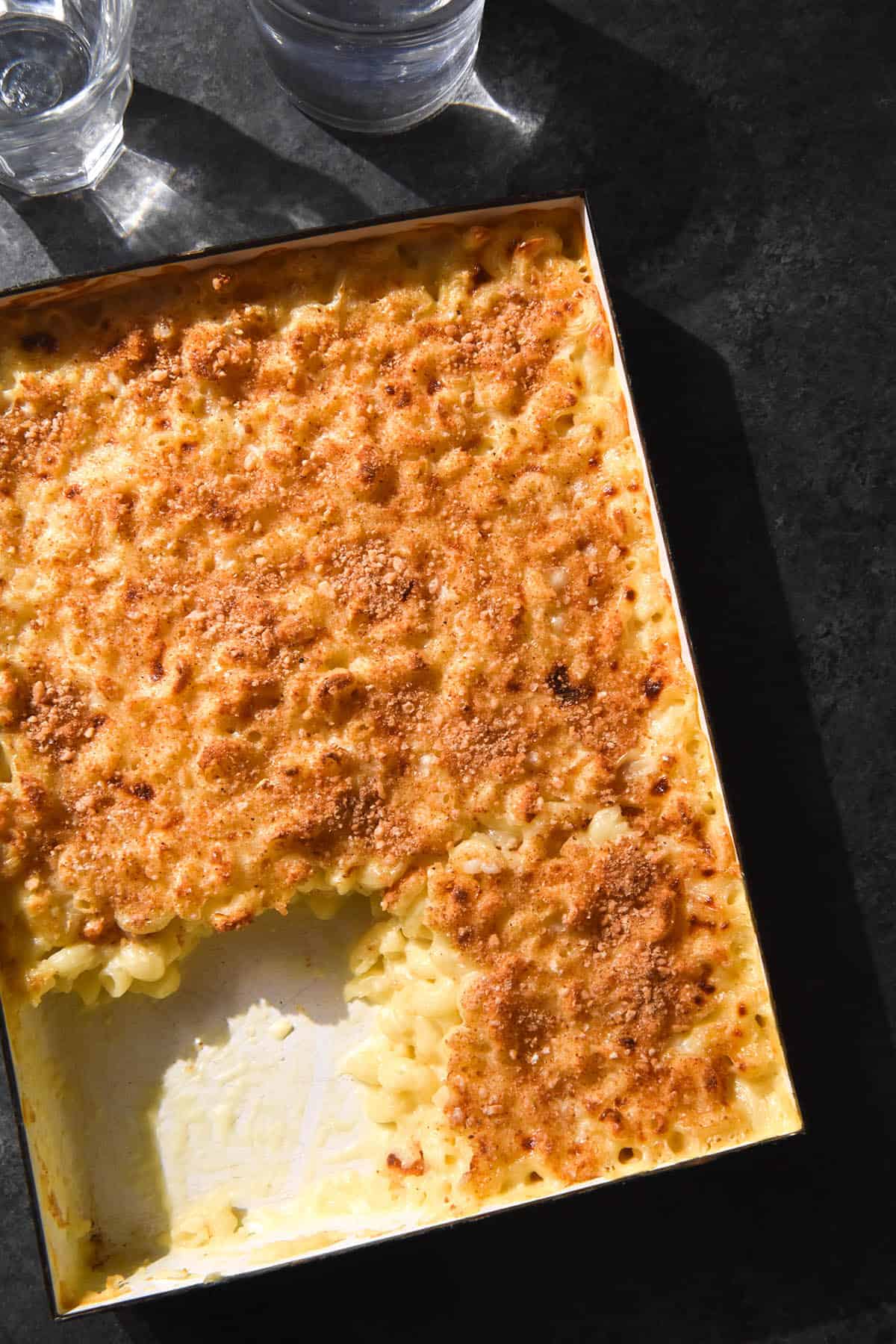 An aerial image of a low FODMAP mac and cheese bake in a white dish atop a dark backdrop. The bottom left piece of the mac and cheese bake has been removed. 