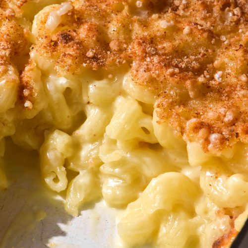 A close up of a mac and cheese bake in a white baking dish. Some of the bake has been removed, revealing the creamy mac and cheese underneath the golden brown topping.