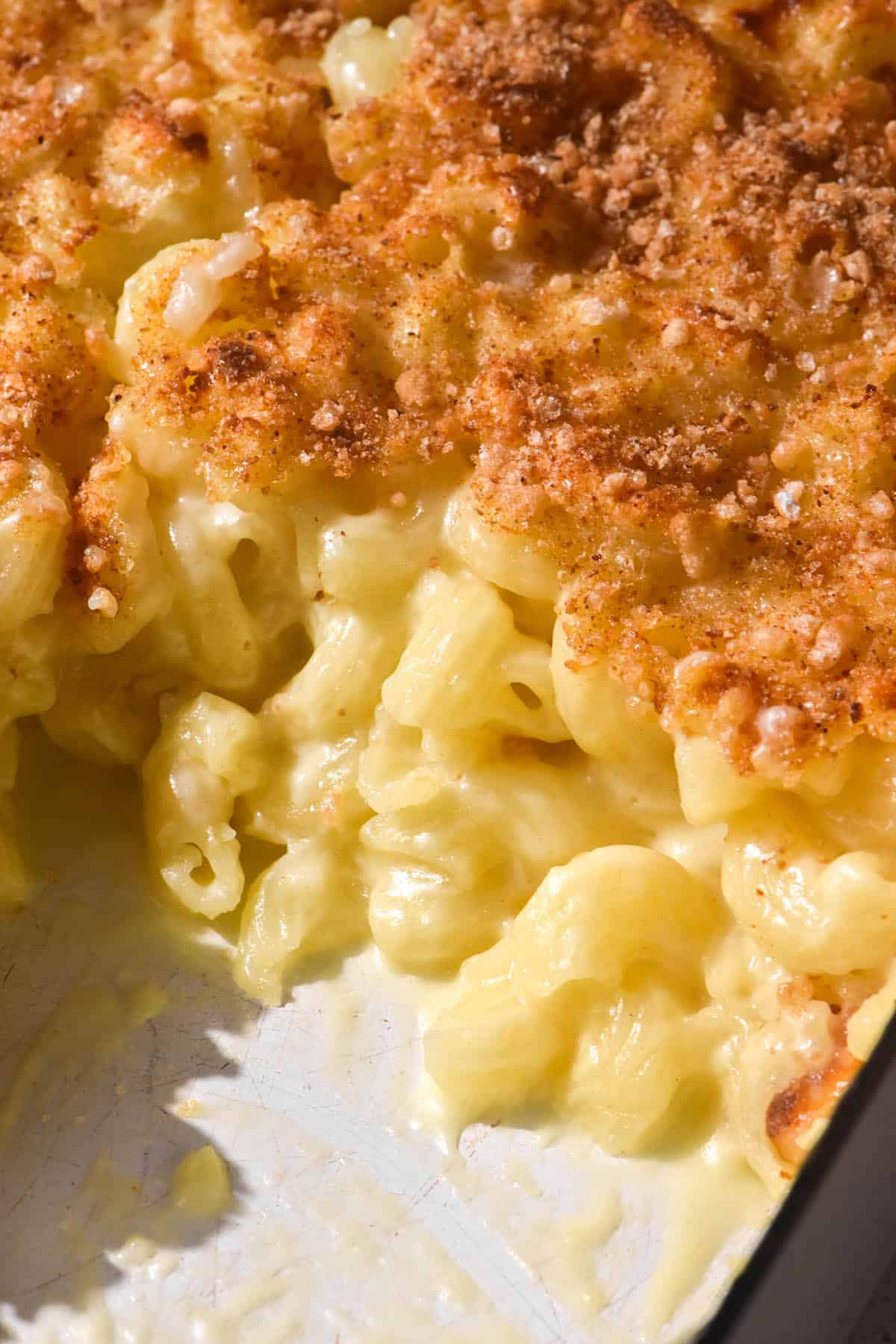 A close up of a mac and cheese bake in a white baking dish. Some of the bake has been removed, revealing the creamy mac and cheese underneath the golden brown topping.