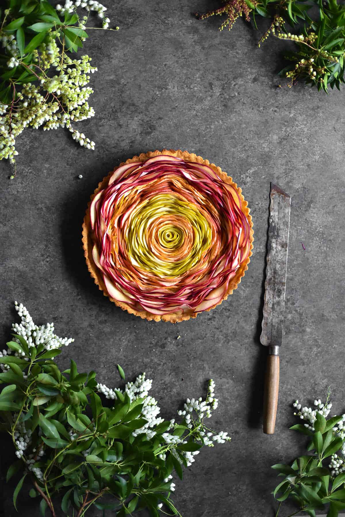 An aerial image of a swirled apple tart on a mottled grey backdrop. The tart is surrounded by floral greenery and sat next to a vintage knife. 
