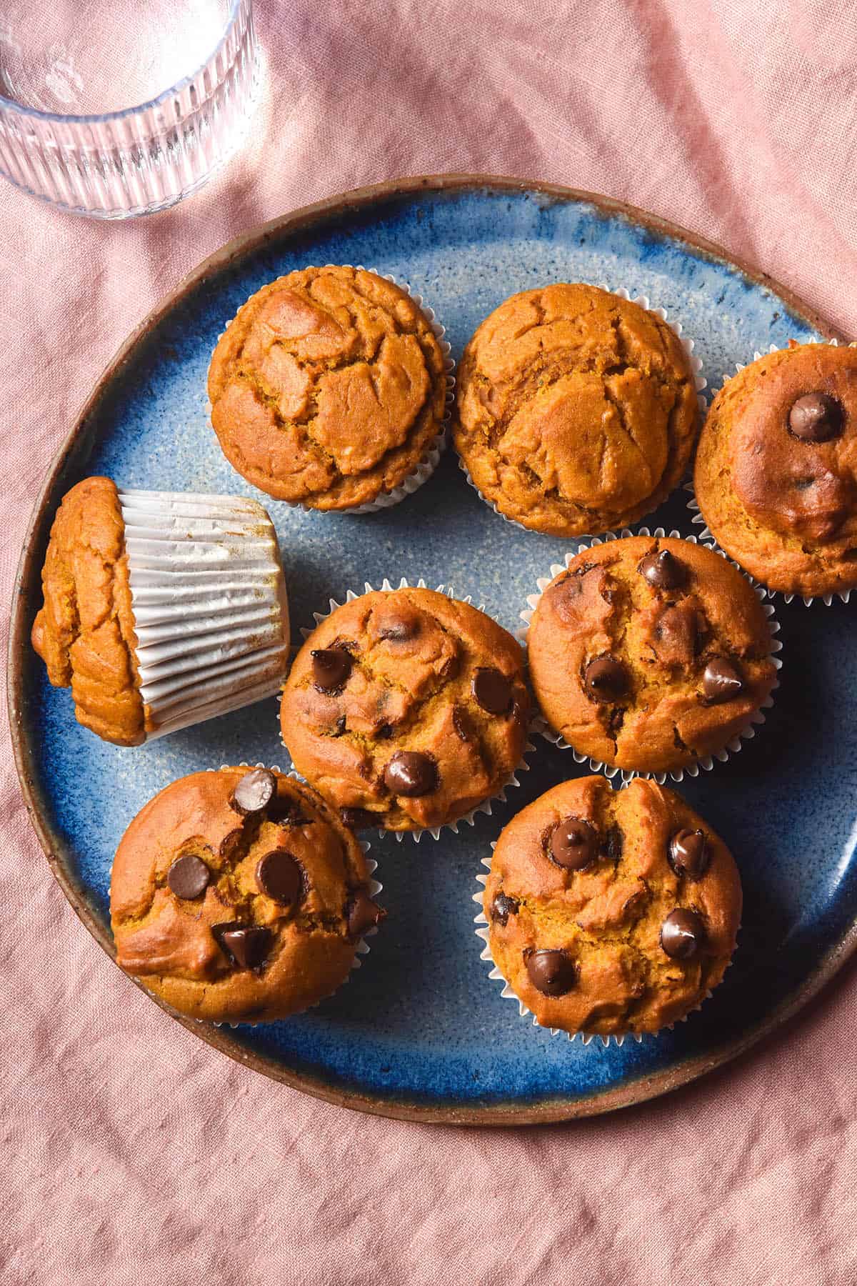 An aerial image of gluten free pumpkin muffins on a bright blue ceramic plate atop a pale pink linen tablecloth. Some of the muffins are studded with melty dark chocolate chips. 