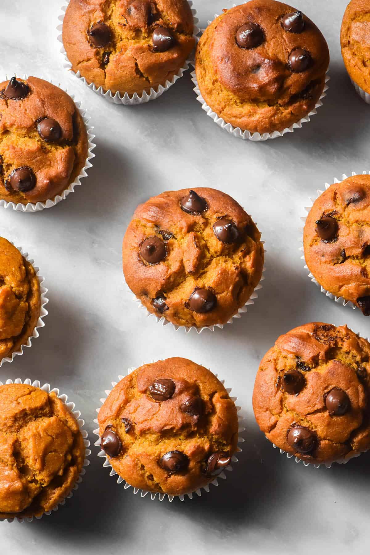 An aerial image of gluten free pumpkin muffins topped with choc chips arranged casually on a white marble table. 