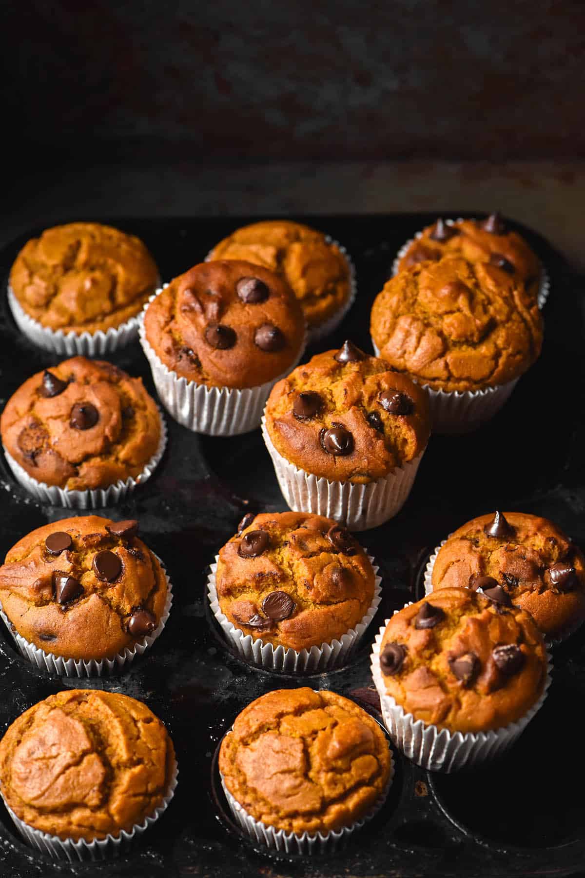 A dark and moody image of gluten free pumpkin choc chip muffins in white muffin liners casually arranged on a dark mottled muffin tray against a black backdrop.
