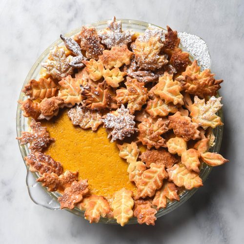 An aerial image of a gluten free pumpkin pie decorated with pastry leaves on a white marble table.
