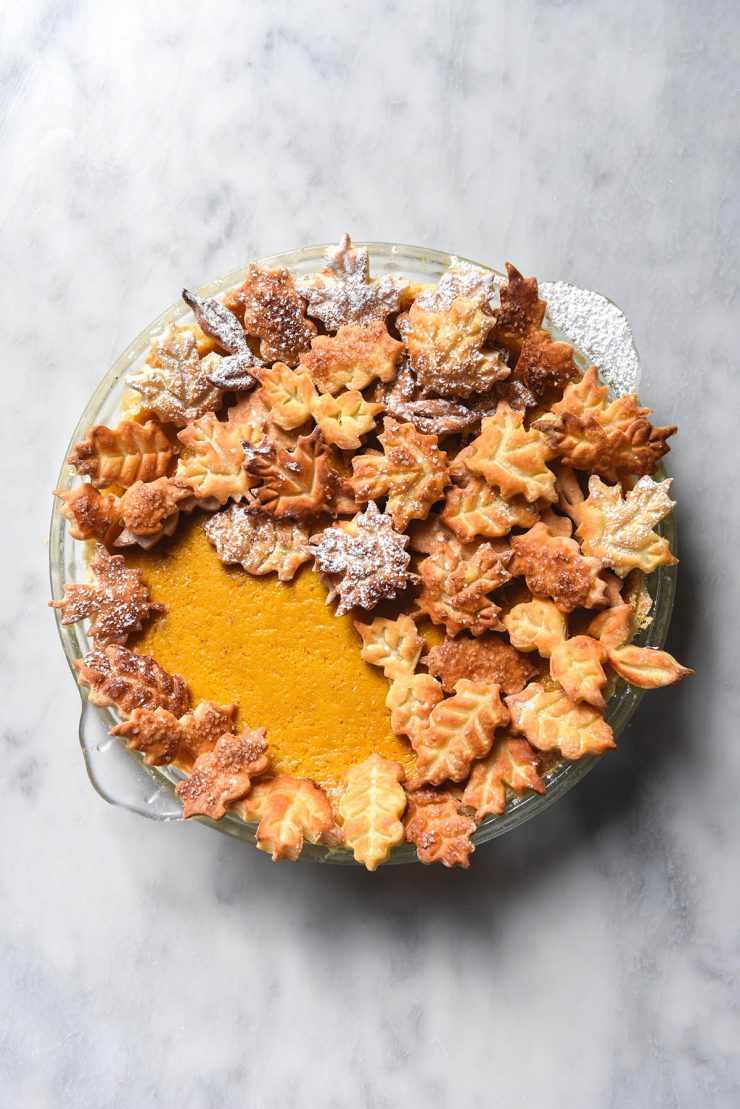 An aerial image of a gluten free pumpkin pie decorated with pastry leaves on a white marble table.