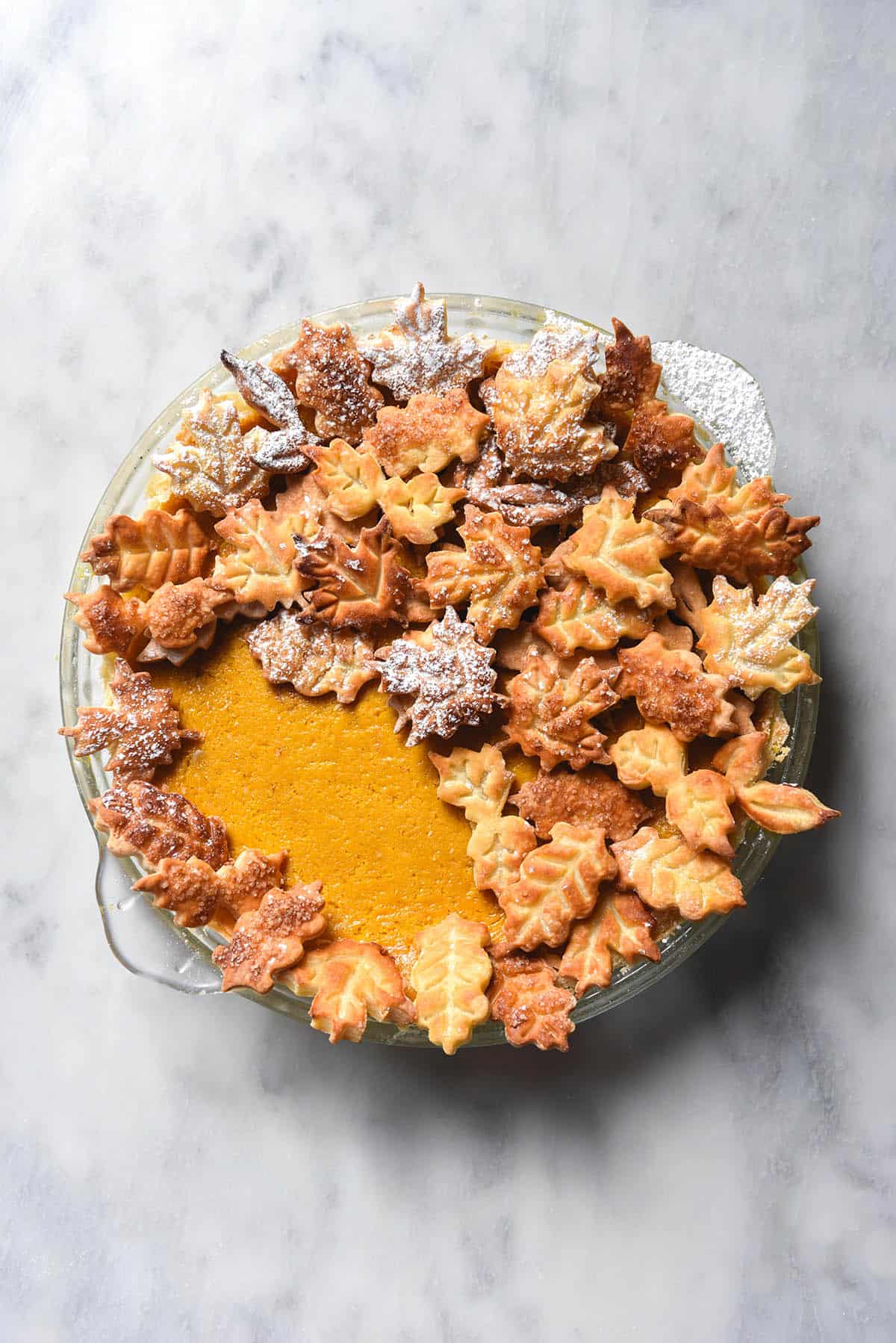 An aerial image of a low FODMAP pumpkin pie decorated with pastry leaves on a white marble table.