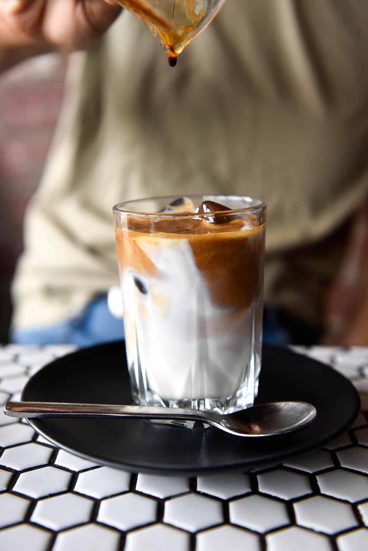 A side on image of an iced latte being poured into a glass cup. A person in an olive green t-shirt sits behind the coffee to pour the espresso into the cup. 