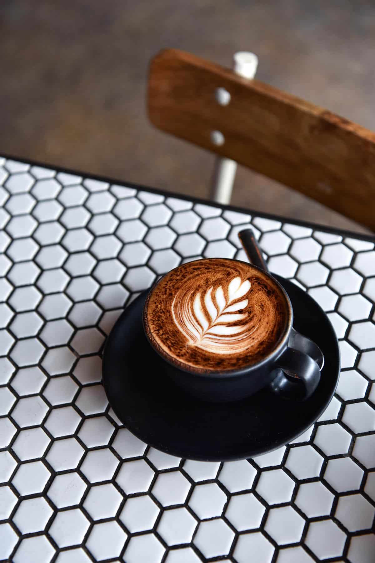 A side on image of cappuccino in a black matte cup and saucer on a white hexagonal tiled table